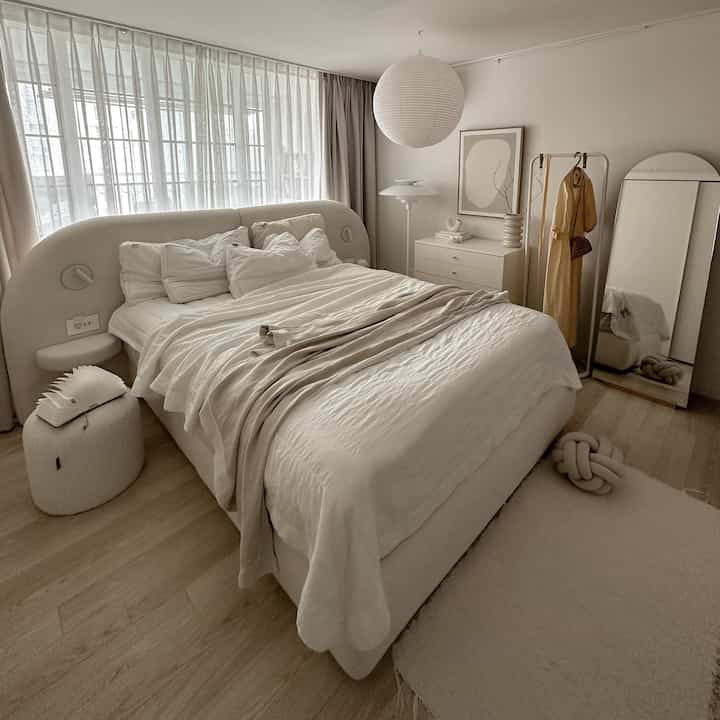 明るいアイボリーとベージュのトーンの寝室、丸みを帯びたフレームベッドとカーテン、全身鏡が配置された雰囲気のあるインテリア