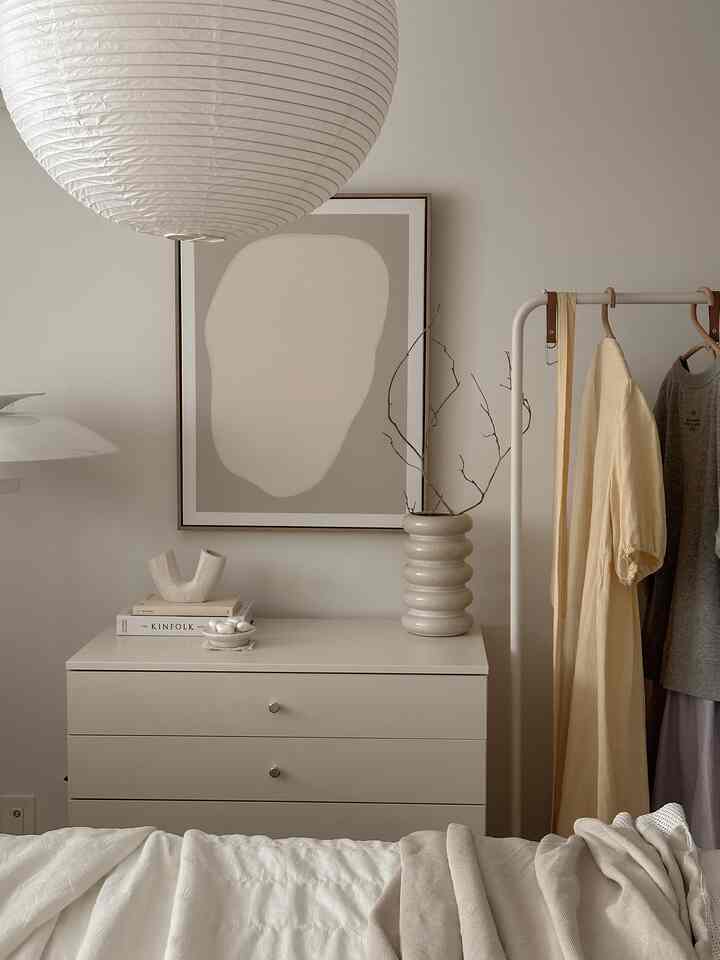 White and beige toned bedroom featuring a dresser with objet and picture frame, and a coat rack on the right in a minimal natural style
