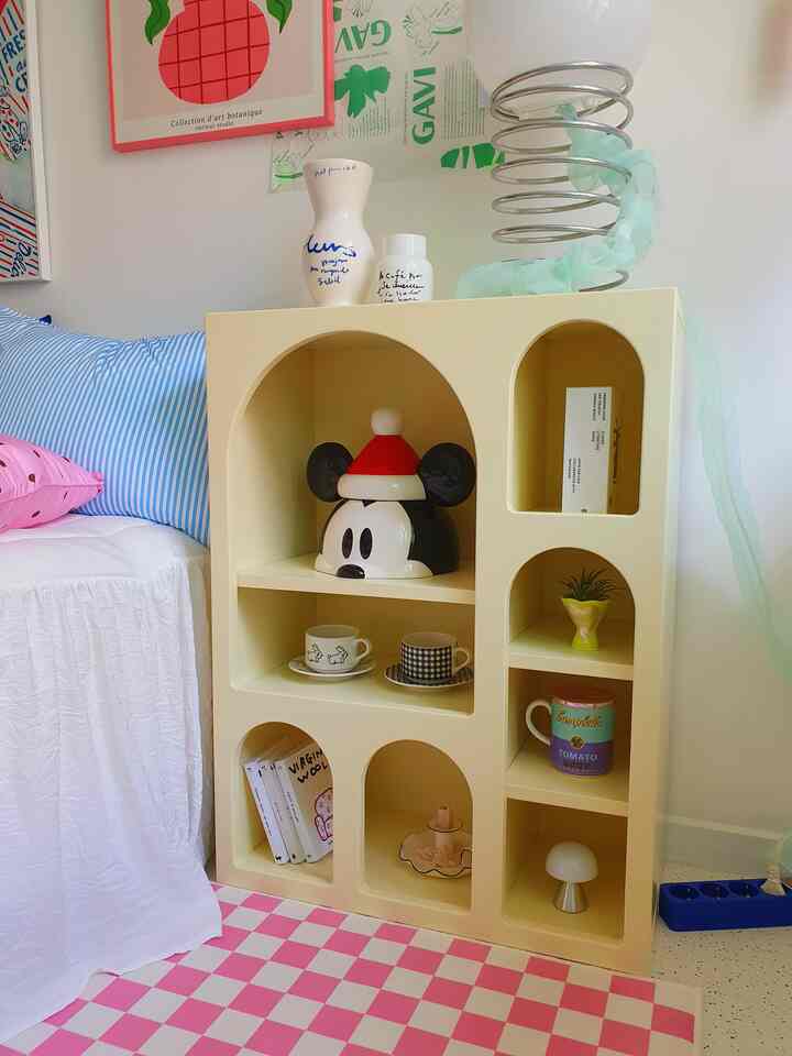 Pastel yellow arched shelf cabinet in a bedroom-like living space featuring various objets and a pink checkered carpet creating a cozy vibe