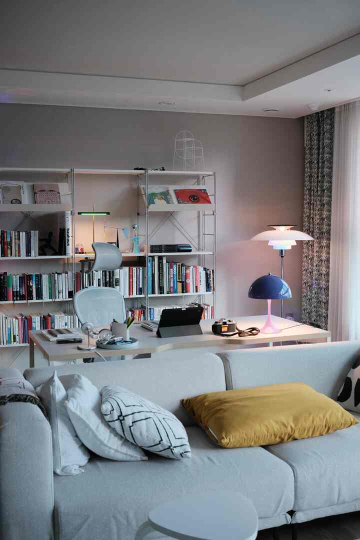 Bright, neutral-toned long living room featuring a sofa, Aeron chair behind desk, large bookshelf, and floor lamp creating a cozy, functional home office.