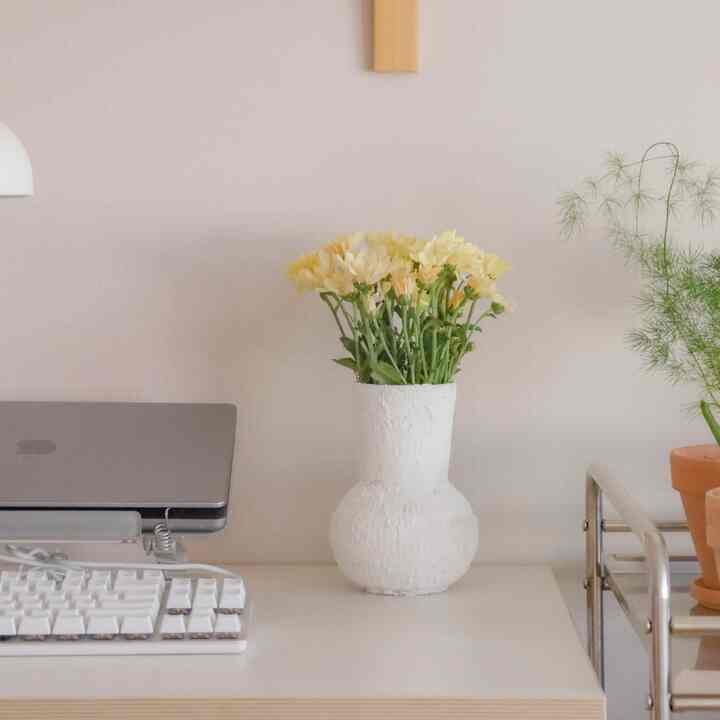 White and brown toned studio home office featuring a Desker desk, IKEA lighting, vase with flowers, and plants creating a warm atmosphere