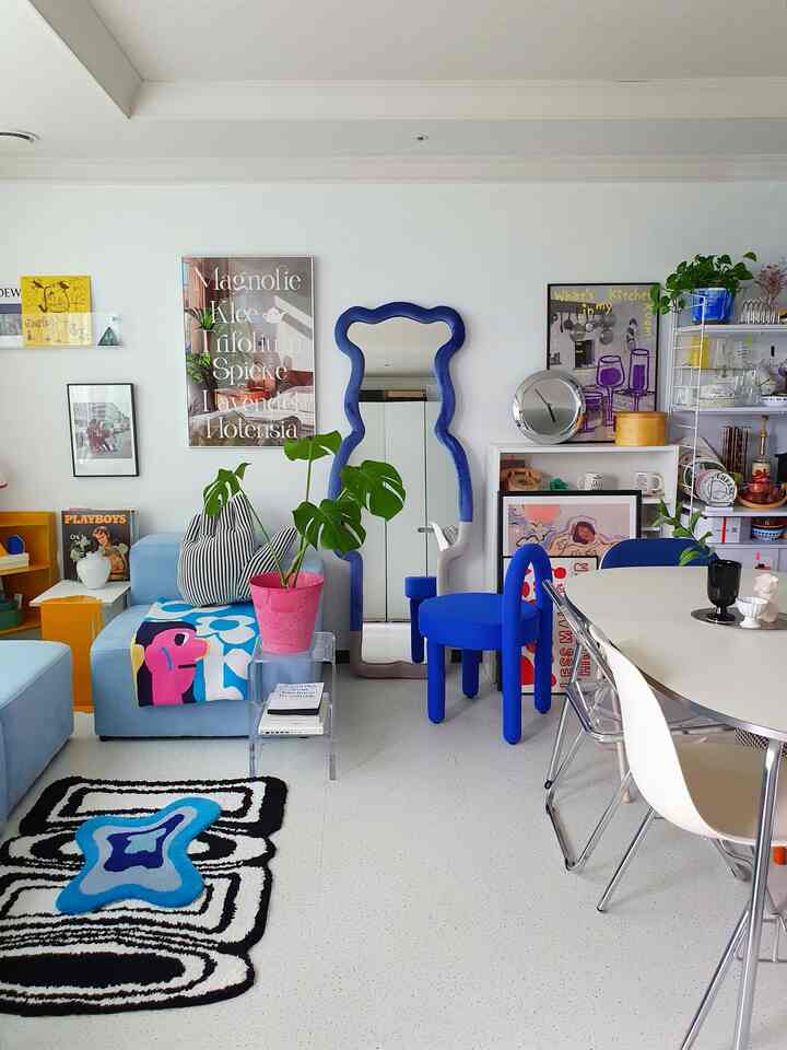 白い壁と床のモダンなリビング、青い全身鏡と青いスツール、多彩なソファと額縁が配された空間