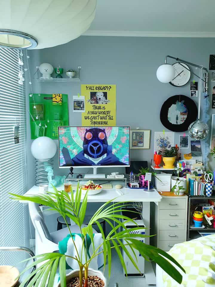 Gray-walled home office with white desk and monitor, featuring green plants in the foreground creating a cozy workspace