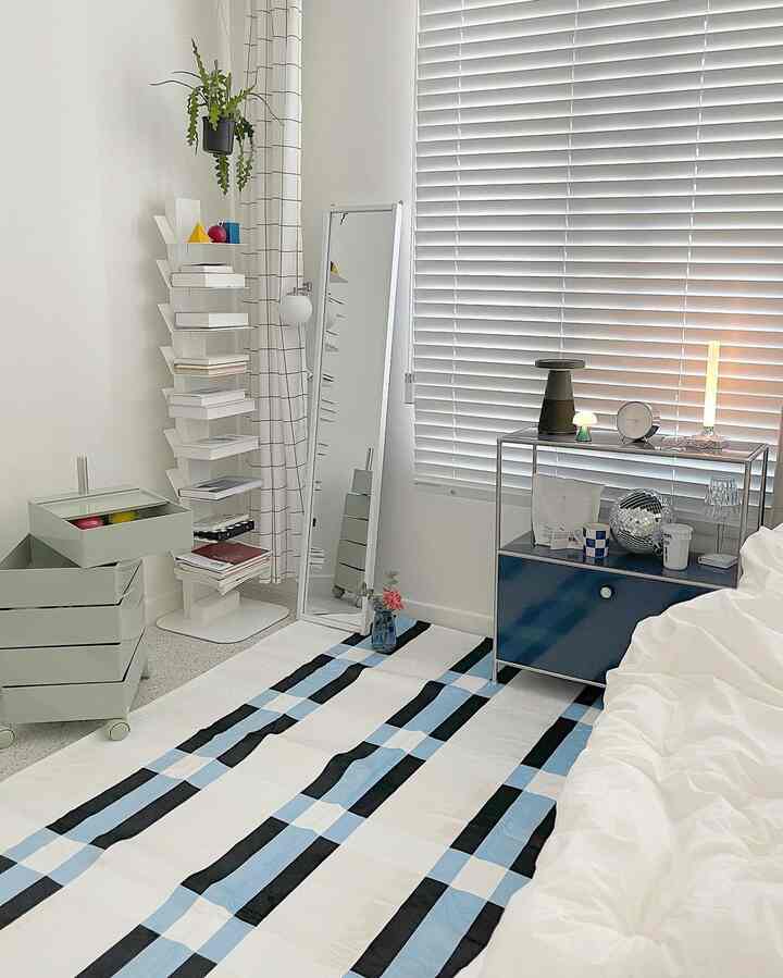 ホワイトとブルーのトーンが調和したリビング、チェック柄ラグと本棚、姿見、トロリーが配置された清潔感のあるモダンなインテリア