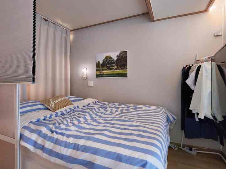 アイボリーとブルーの色調の寝室、リフトアップベッドとストライプの寝具、右側にホームウェア用ハンガーが配置された実用的なインテリア