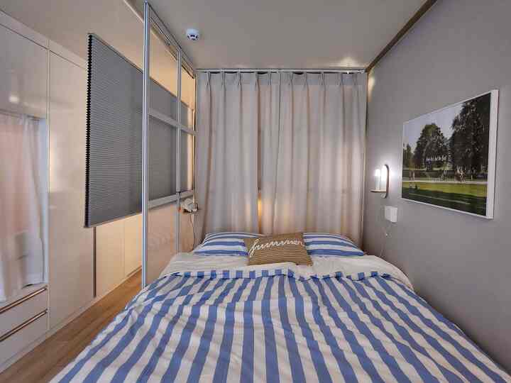 Modern studio bedroom with white walls and blue-striped bedding, featuring room dividers and honeycomb blinds creating practical separated spaces