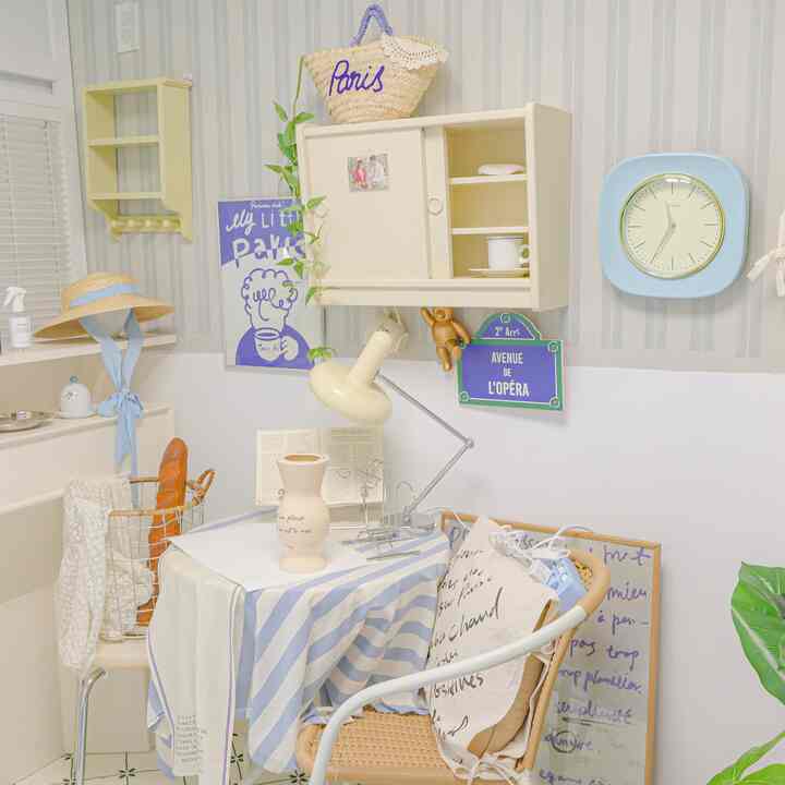 柔らかなブルーとアイボリーの色調の寝室、テーブルランプや壁時計、小さなかわいらしいインテリアが配置された雰囲気のある空間