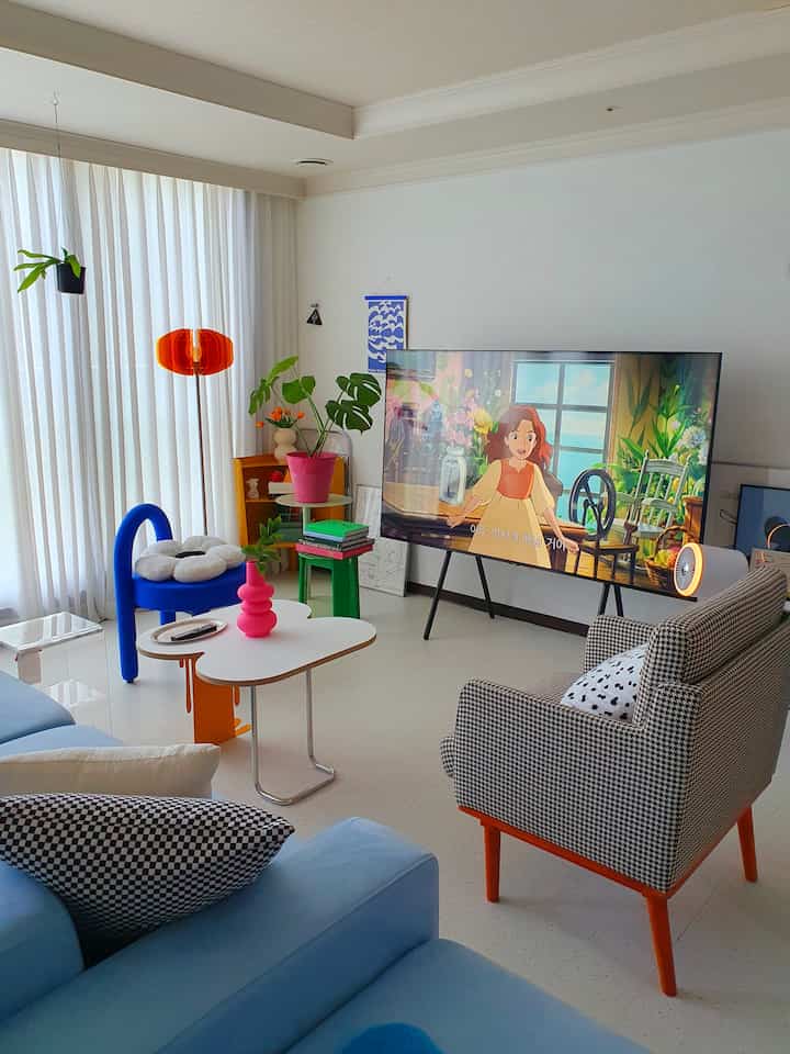 Bright living room dominated by white and orange accents featuring mid-century modern sofa and armchair, large TV, and lush plants creating vibrant ambiance