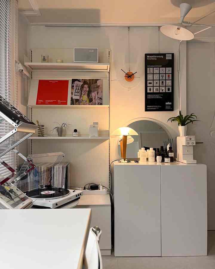 White-toned home office featuring wall shelves, a cabinet with a turntable and mirror, highlighted by warm lighting in a tidy interior