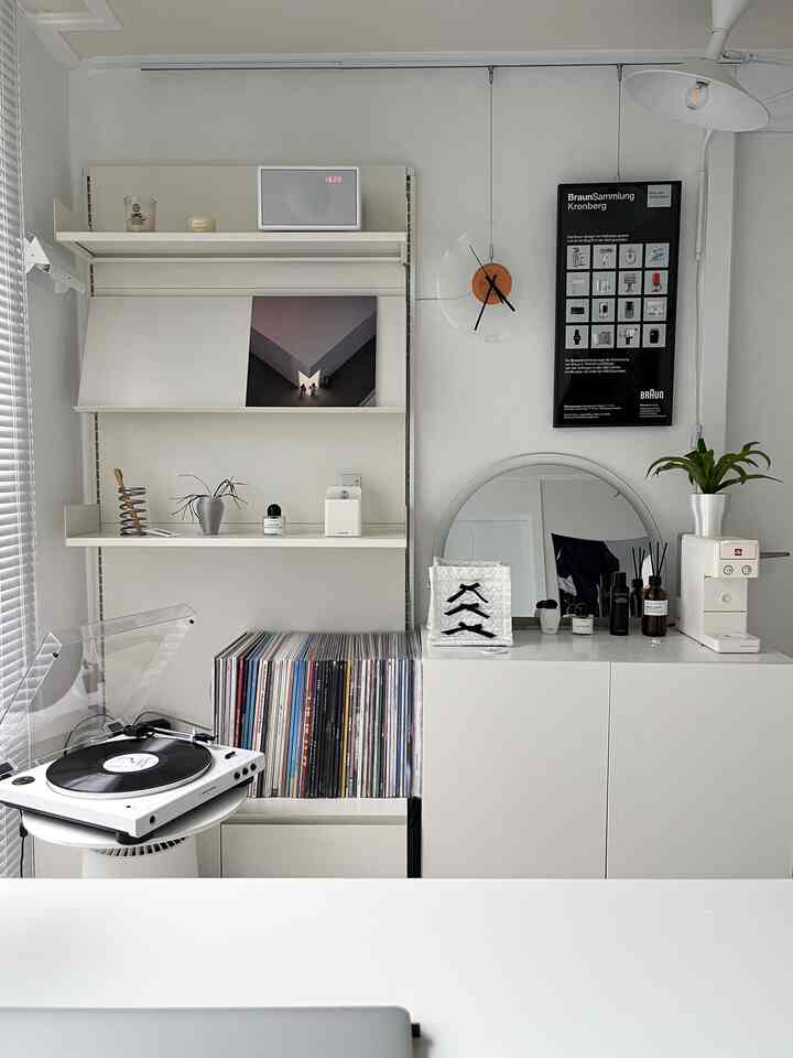 White-toned home office space featuring wall shelves, turntable and vinyl LP collection, round mirror and lighting in modern interior