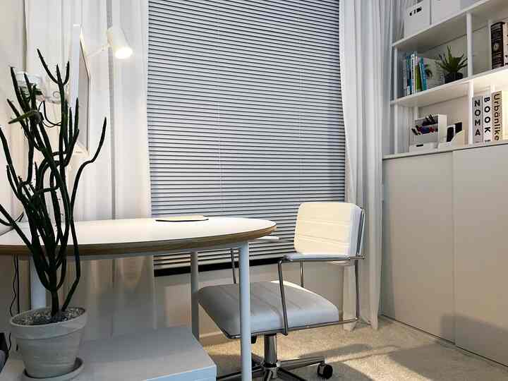 白とナチュラルトーンのホームオフィス、白いデスクとチェア、大きなグレーのブラインド、本棚と植物が配置された清潔で落ち着いたインテリア