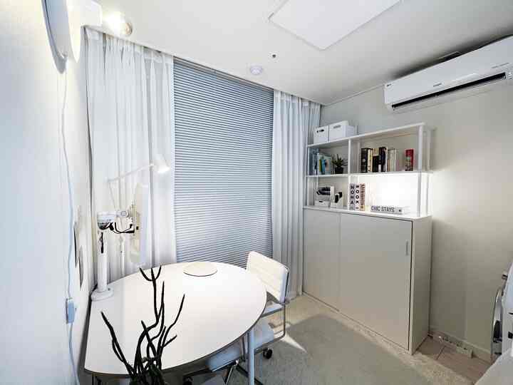 ホワイトトーンのホームオフィス空間、丸テーブルと本棚、シフォンカーテンとブラインドが配置された温かみのある部屋