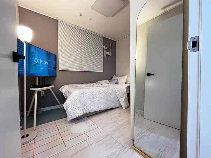 グレーとホワイトのトーンのワンルーム寝室、シングルベッド、テレビ、全身鏡、フロアランプが配置された清潔でモダンな空間