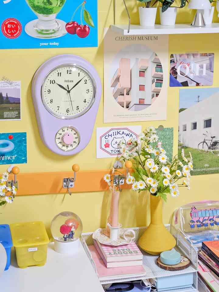 A bright yellow wall with white desk, lavender wall clock, and yellow vase with flowers create a charming Korean interior kidult room