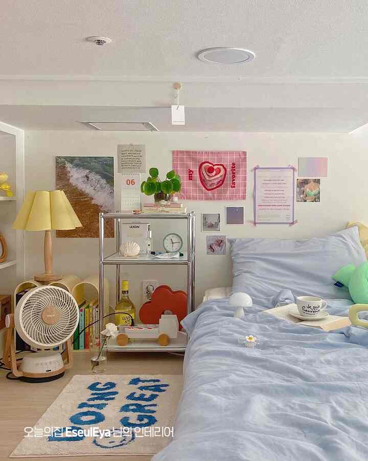 白い壁と青い寝具が映えるワンルームの寝室で、ミッドセンチュリーモダン風のメタルシェルフと黄色いテーブルランプが配置された雰囲気のあるインテリア。