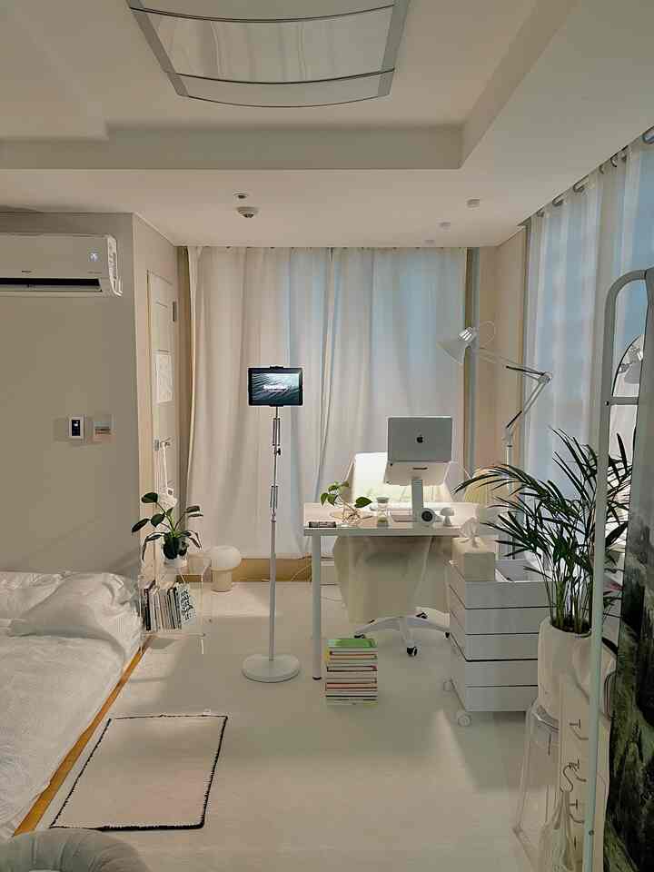 明るいホワイトトーンの1人用寝室兼ホームオフィス、机とコンピューター、照明が配置された清潔感あるインテリア