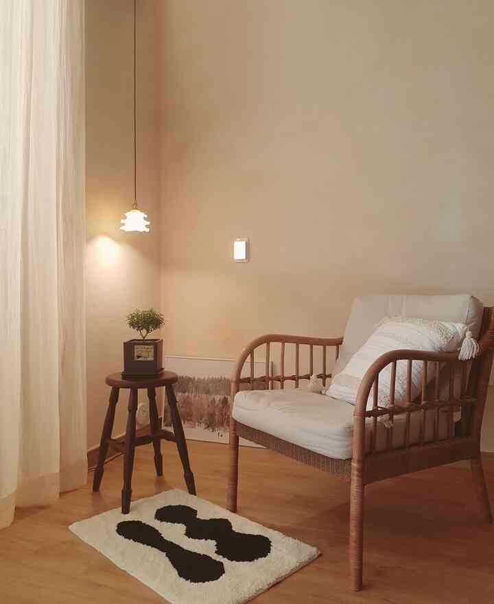Beige-walled studio living room with wood-tone flooring, featuring a rattan armchair, stool, curtain, and plant in a cozy setup