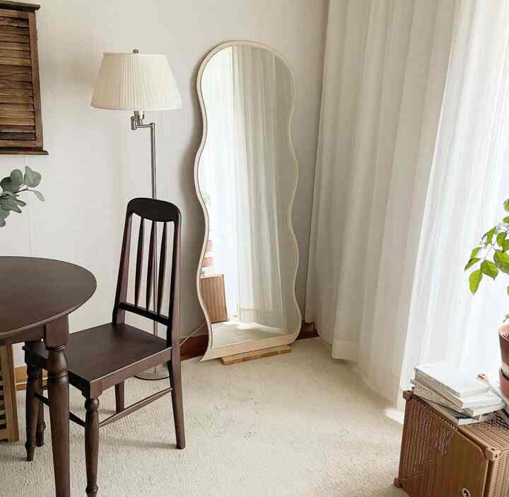 アイボリーの色調のリビング、ヴィンテージの木製ダイニングテーブルと椅子、波形の全身鏡、白いフロアランプが配置された自然な雰囲気のインテリア