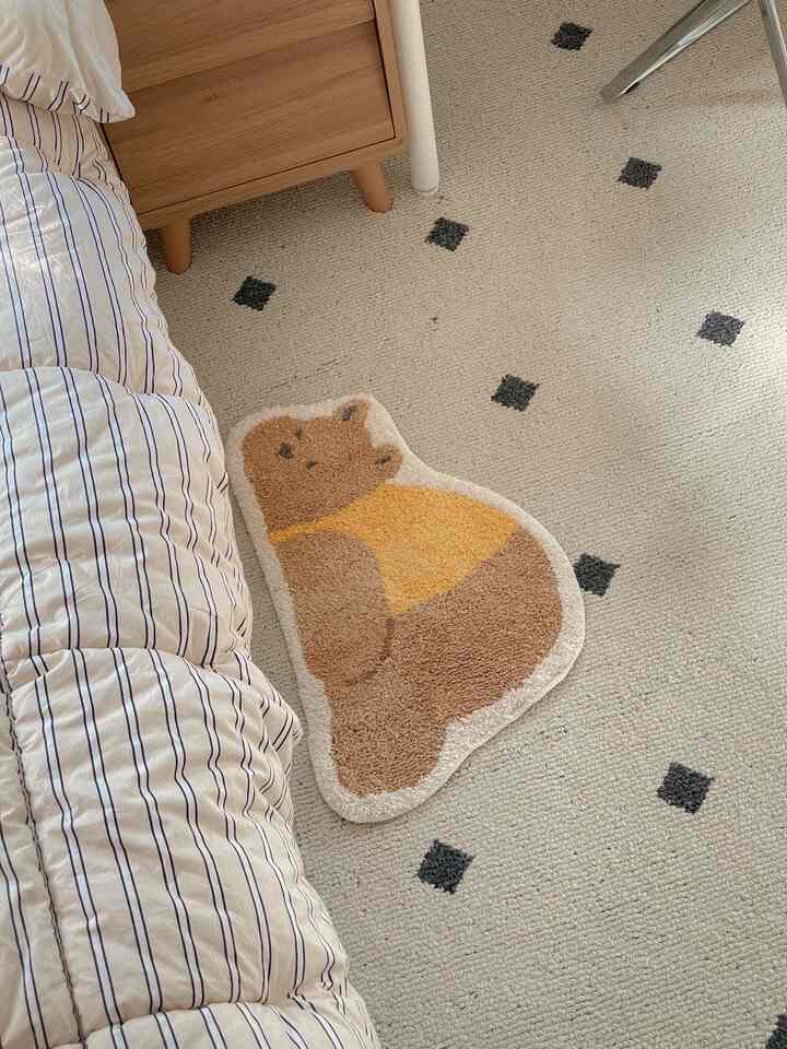 ベージュ色の床にかわいいクマの形のポイントラグが置かれた寝室の一部、ストライプの寝具と木製のサイドテーブルが温かみのある雰囲気を演出する空間