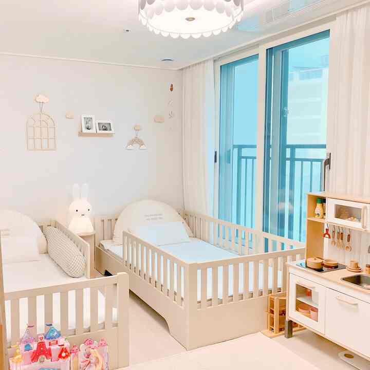 Bright and clean kids' room with white and wood tones featuring two cribs and a wooden play kitchen set