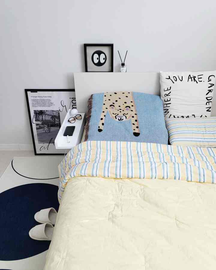 白い壁と木目調のナイトテーブルがある温かみのある寝室、黄色ベージュと青のストライプ寝具が雰囲気を演出