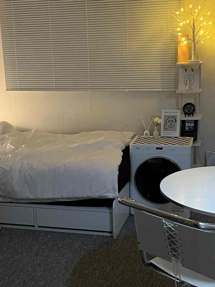 White and gray toned studio apartment bedroom featuring bed, washing machine, and bookshelf arranged neatly in cozy space