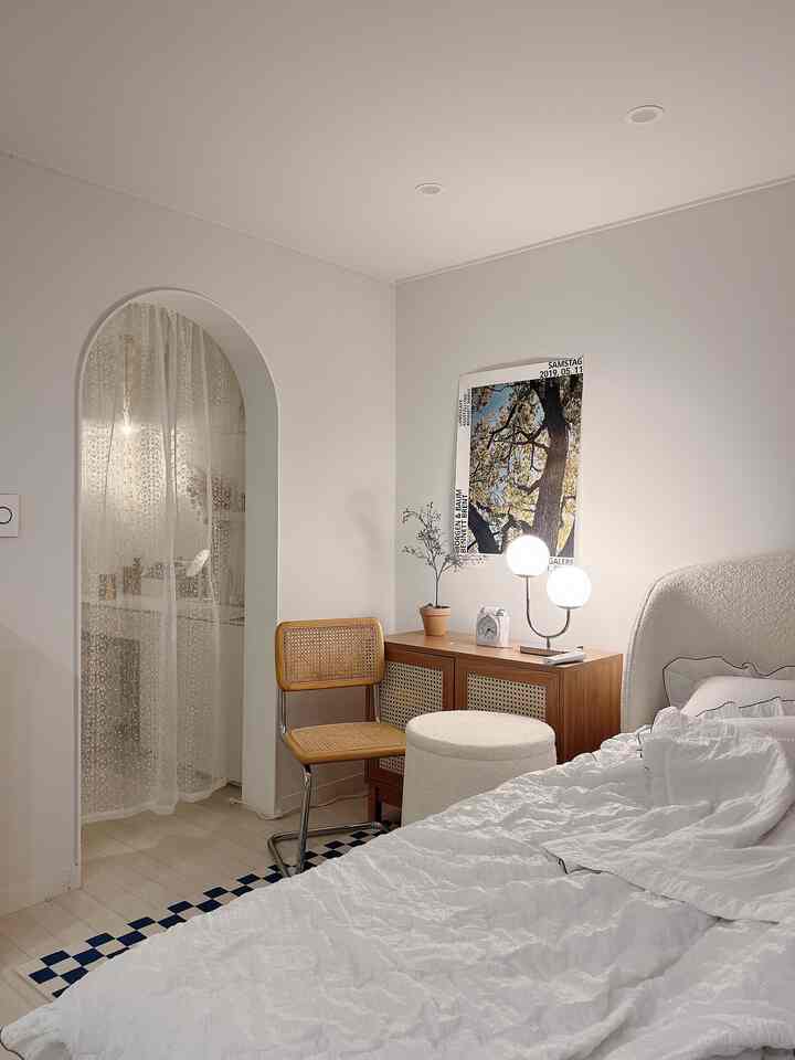 A cozy bedroom in white and beige tones featuring an arched entrance with lace room divider curtain, rattan chair, and cabinet.