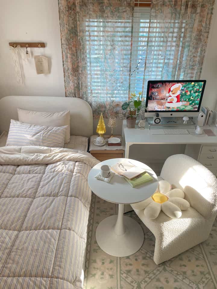A white and beige toned bedroom featuring a clean desk, bed, round table, and plush armchair with a cozy atmosphere