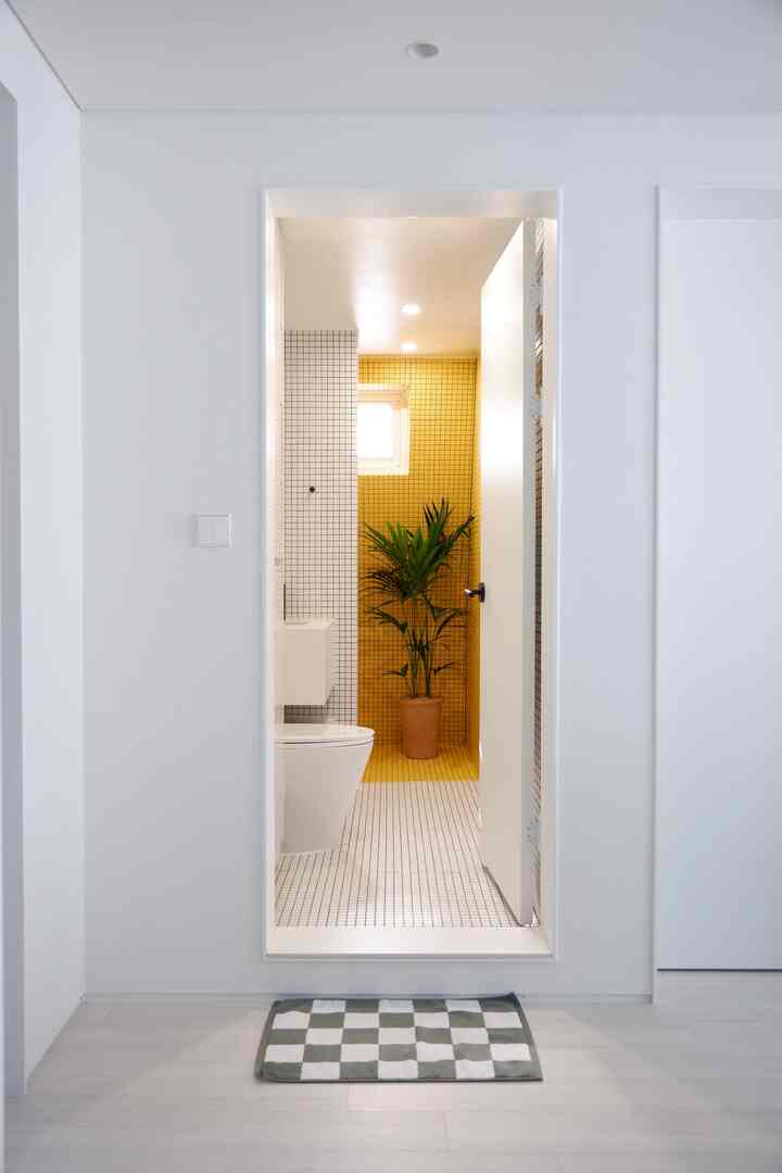 白と黄色のタイルが印象的な狭いモダントイレ、観葉植物とチェック柄のバスマットが配置された雰囲気ある空間