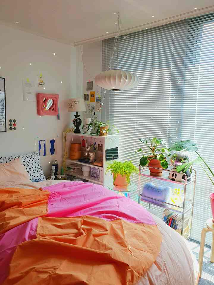 明るいライトグレーの寝室、ピンクとオレンジの寝具、多彩な植物、ミッドセンチュリーモダンの照明がある雰囲気あるインテリア