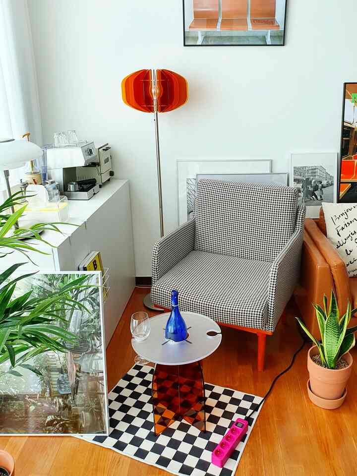 Modern living room with white walls and wooden flooring, featuring a houndstooth armchair, orange floor lamp, and acrylic side table