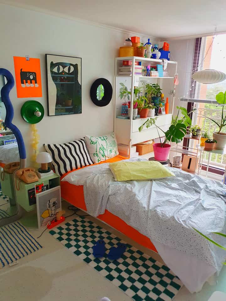 White and orange toned studio bedroom featuring a bed, diverse plants, and posters creating a cozy and vibrant atmosphere