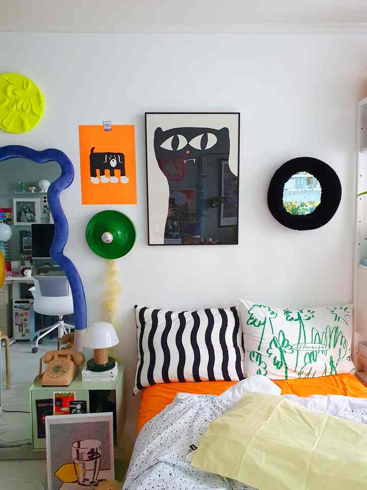 白い壁とオレンジの寝具が印象的なワンルーム寝室、個性的なポスターと曲線状の鏡が調和したモダンでカラフルなインテリア
