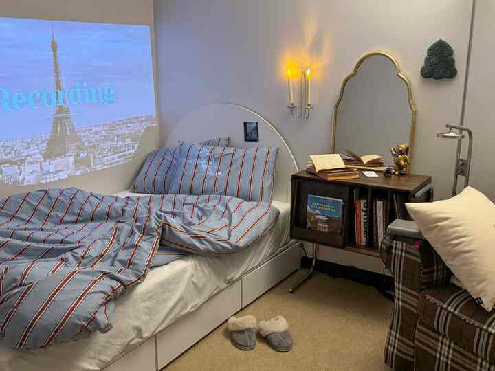 白い壁とベッドにブルーストライプの寝具があり、木製のナイトスタンドとゴールドの鏡が配された雰囲気のある寝室