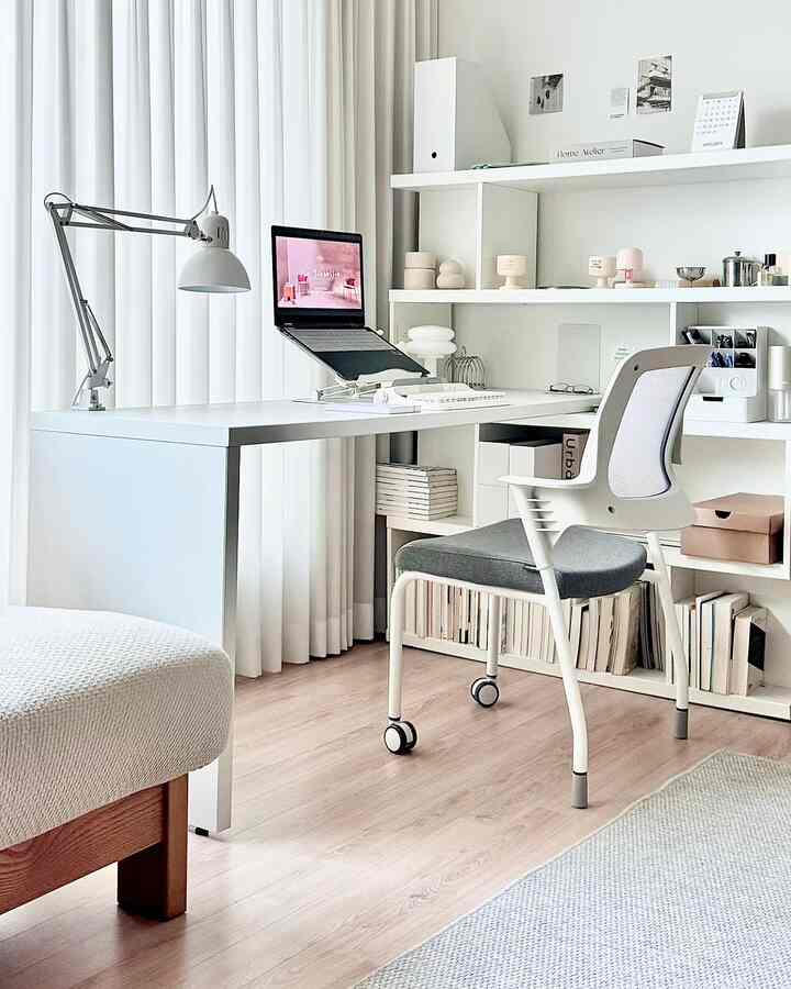 Bright white-toned home office featuring a white desk and chair, laptop stand, and curtains allowing natural light, creating a modern workspace