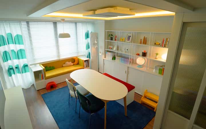 明るくナチュラルな色調のリビング、中央のダイニングテーブル、黄色のソファ、青いラグ、本棚が配置された子供の読書と学習空間