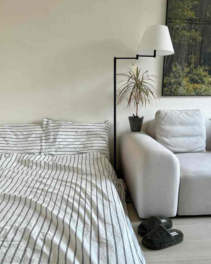 白を基調とした寝室、ストライプの寝具セットとミニマルなソファ、フロアランプが配置された落ち着いた雰囲気のインテリア