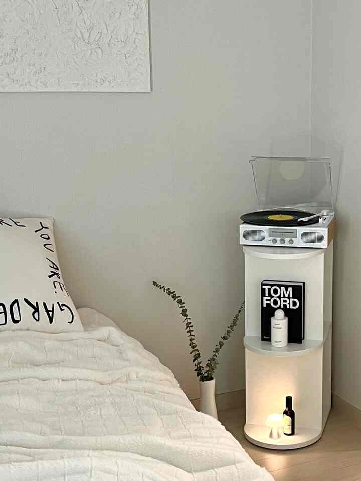 White-toned bedroom featuring clean bedding, modern shelving unit, a plant, and warm lighting creating a cozy atmosphere