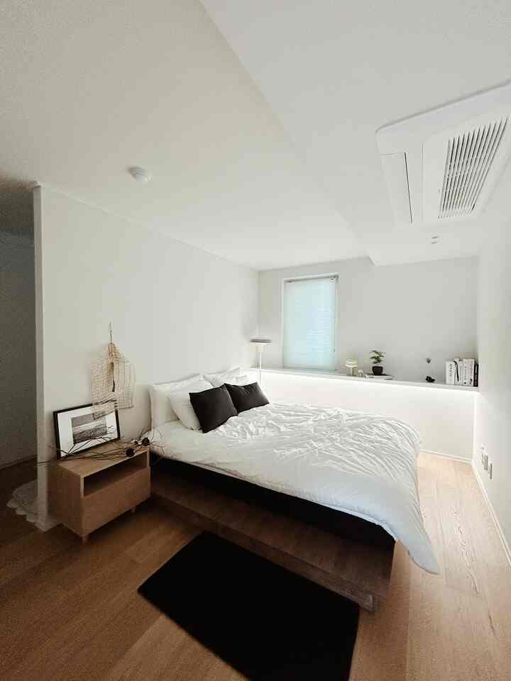 明るいホワイトとブラウンのトーンの広い寝室、中央にベッドと木製ナイトスタンド、窓辺の棚には植物と装飾品が配置された雰囲気のある空間