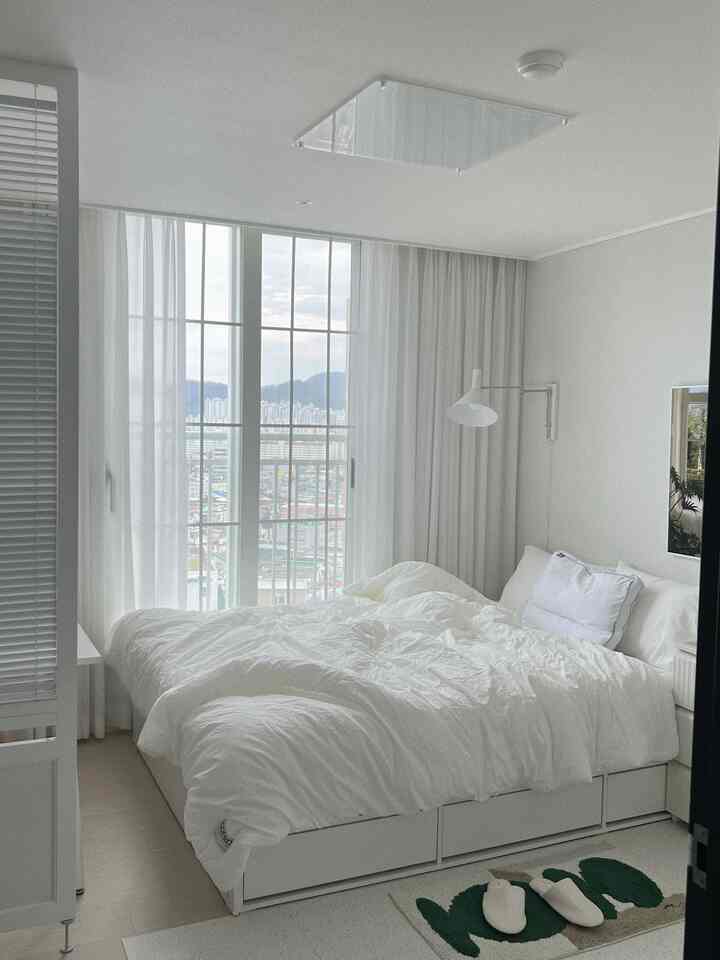 Bright white-toned bedroom with large windows, white bedding, and a wall-mounted lamp creating a clean atmosphere