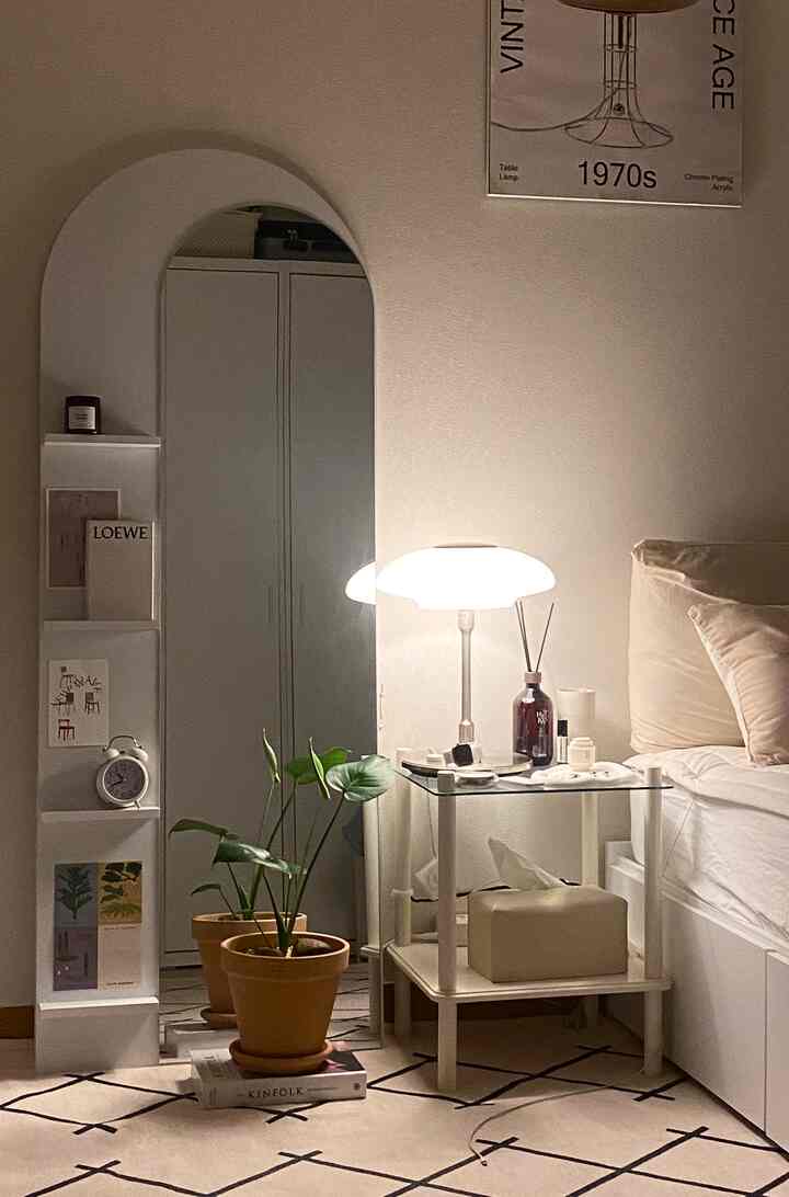 ベージュとモノトーンの寝室、全身鏡とテーブルランプ付きナイトテーブル、テラコッタの植物が配置された落ち着いた印象のインテリア