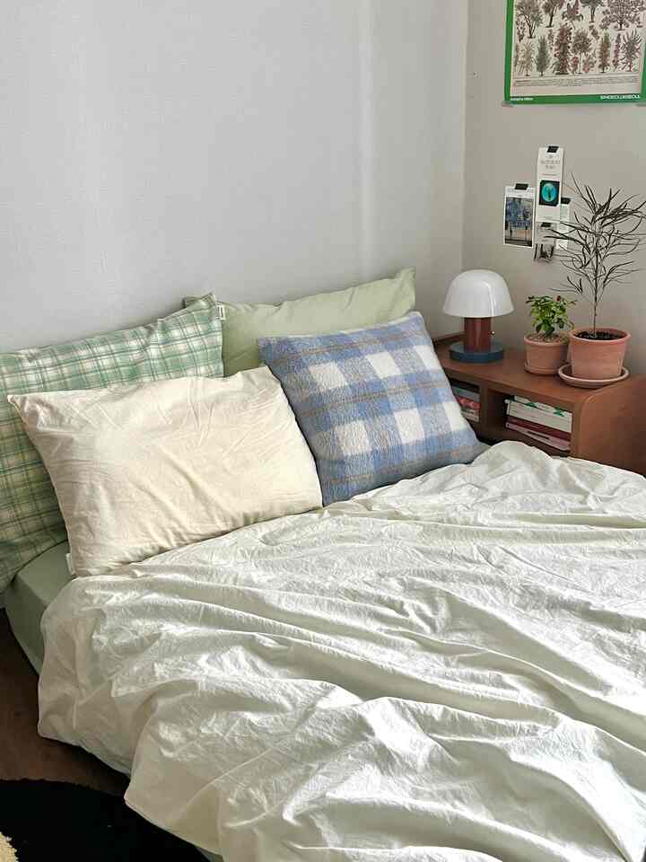 白い壁と寝具のある寝室、ナチュラルな木製ナイトテーブルと緑の植物、パステルカラーのクッションが落ち着いた雰囲気のインテリア