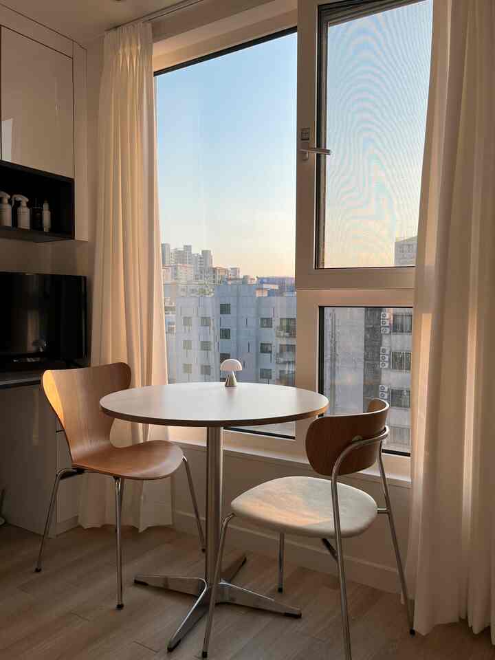 白いカーテンと丸いテーブル、木製の椅子が配置された自然光のたっぷり入る二人用ダイニングスペース