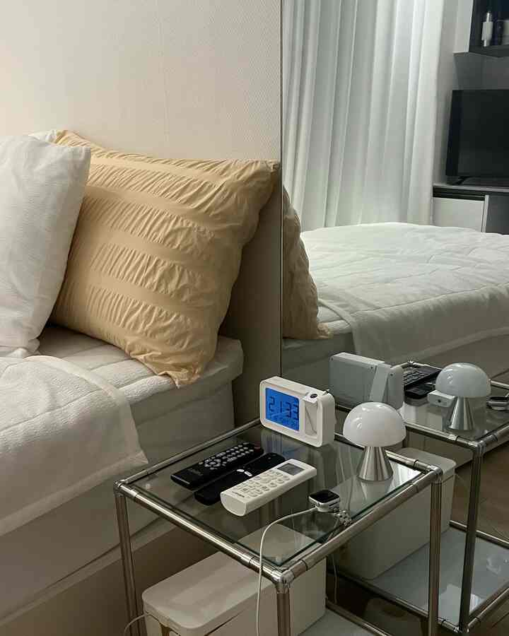 白とベージュの色調の寝室、ナイトテーブルにデジタル時計、リモコン、小さな照明が配置されたミニマルなインテリア