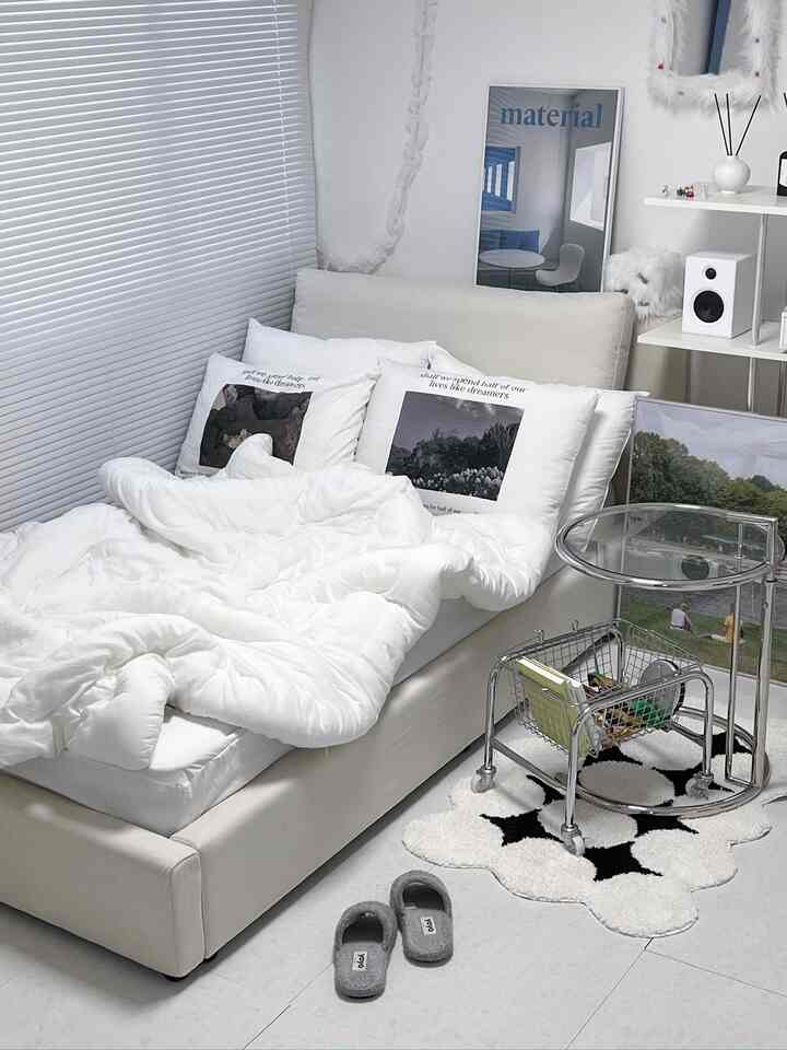 ホワイトトーンの寝室、白いベッドフレームと布団、ブラインド、メタルの収納トロリーが配置されたミニマルで清潔感のあるインテリア