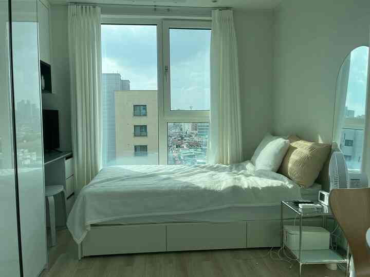 ホワイトとナチュラルトーンのワンルーム寝室、収納可能なベッドフレームと遮光カーテンが配置された清潔感のある空間