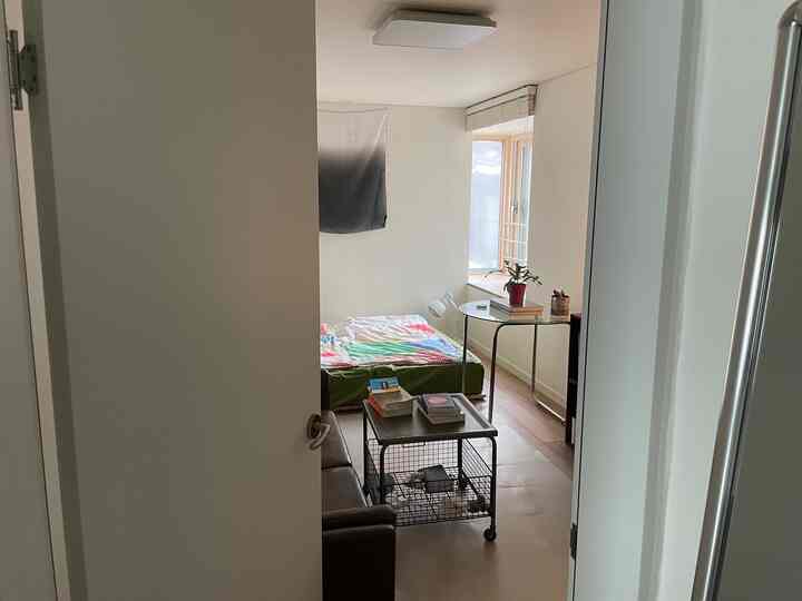 白い壁と茶色のソファ、多彩な寝具がある6畳の小さな部屋、自然光が差し込むガラスの丸テーブル付きのインテリア