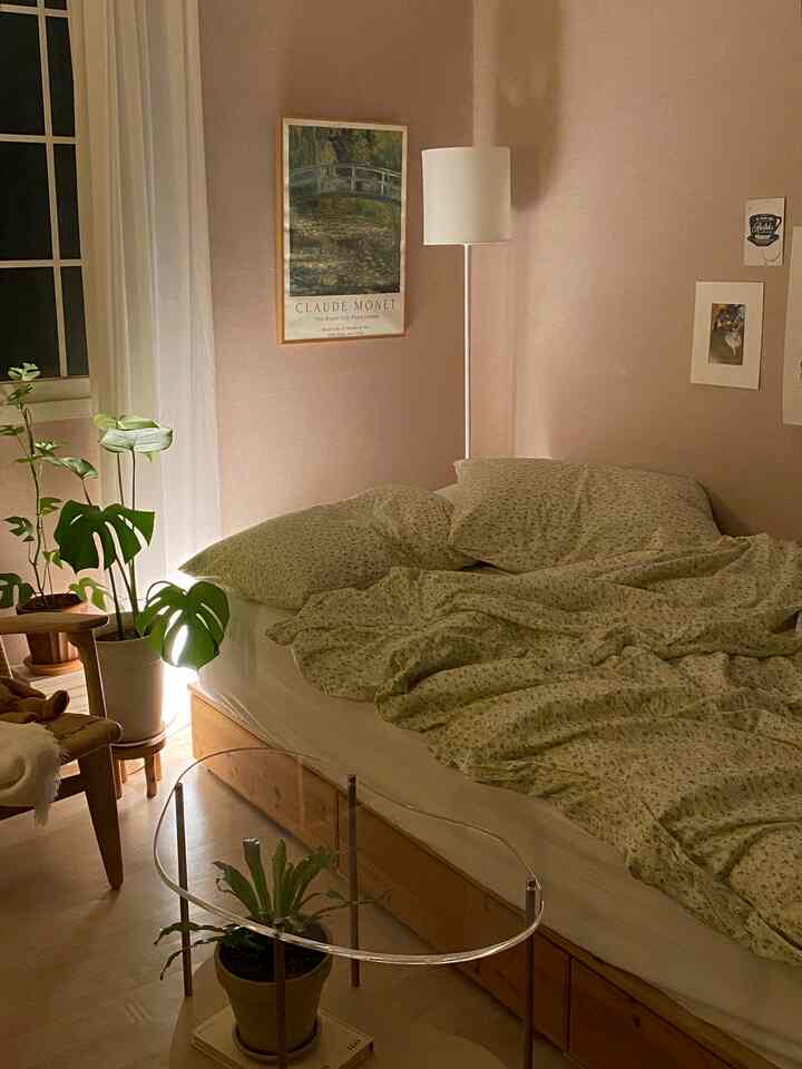 柔らかなピンク色の壁とナチュラルな木目調家具が調和する寝室、ベッド、フロアランプ、植物が落ち着いた雰囲気を作り出す印象的な空間