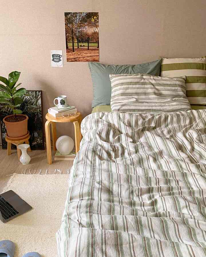 ベージュの壁と木目調の床が調和した寝室で、緑のストライプ柄掛け布団カバーのベッドと木製スツールが落ち着いた雰囲気のインテリア