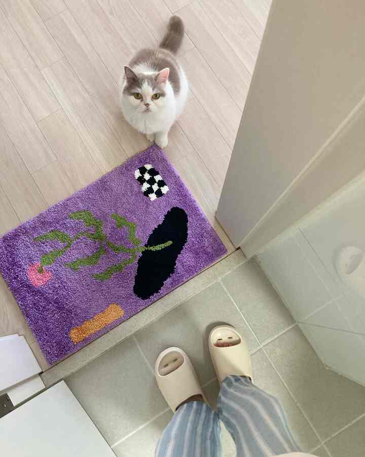 明るいナチュラルカラーの床と白い壁のバスルーム入口、紫のバスマットの上に座る猫とベージュのバススリッパがある空間